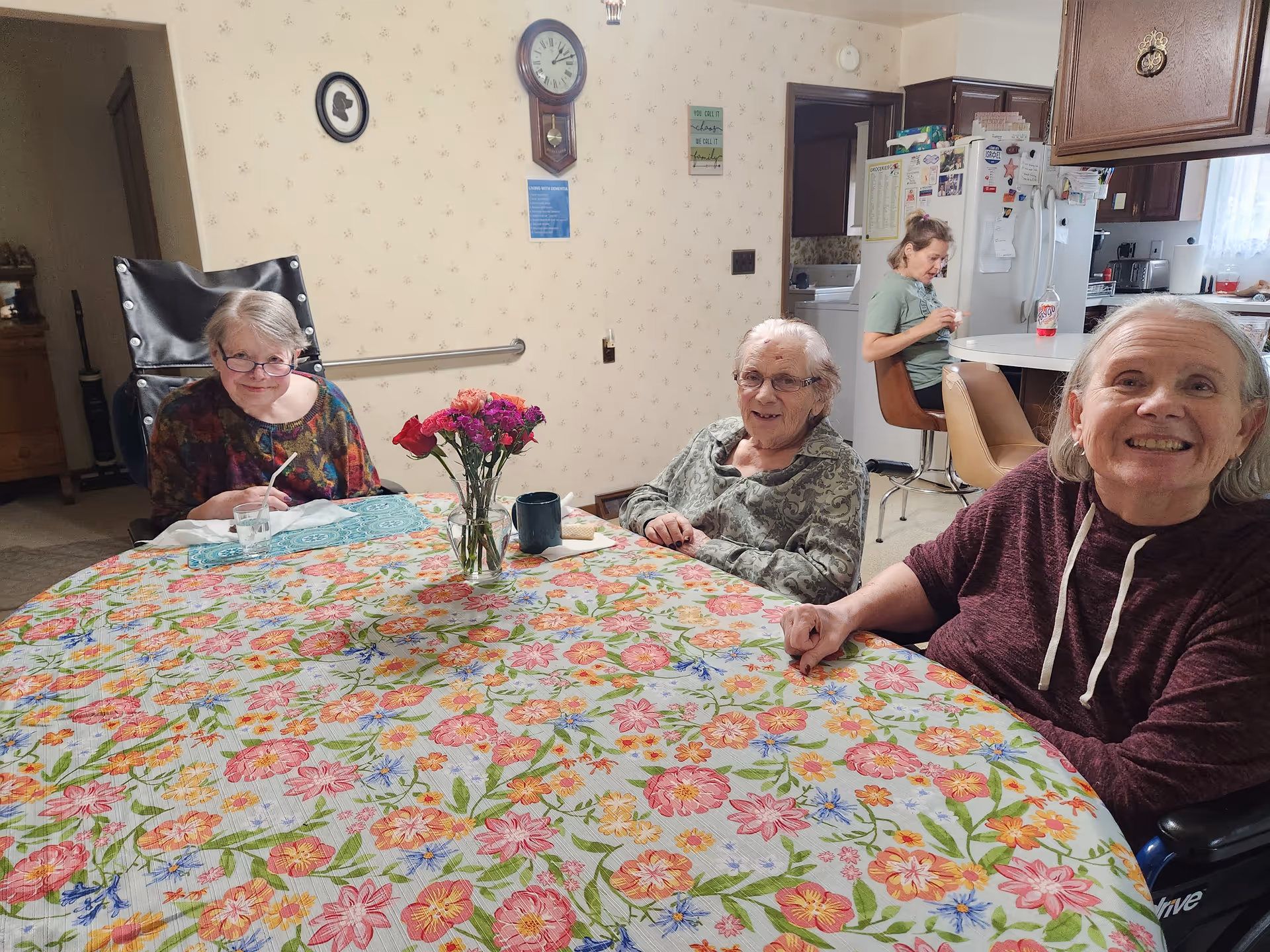 Three elderly women sitting around a round table covered with a colorful floral tablecloth in a cozy room. A vase with flowers is placed in the center of the table. In the background, a younger woman is sitting on a stool near a kitchen counter with a refrigerator and cabinets visible.