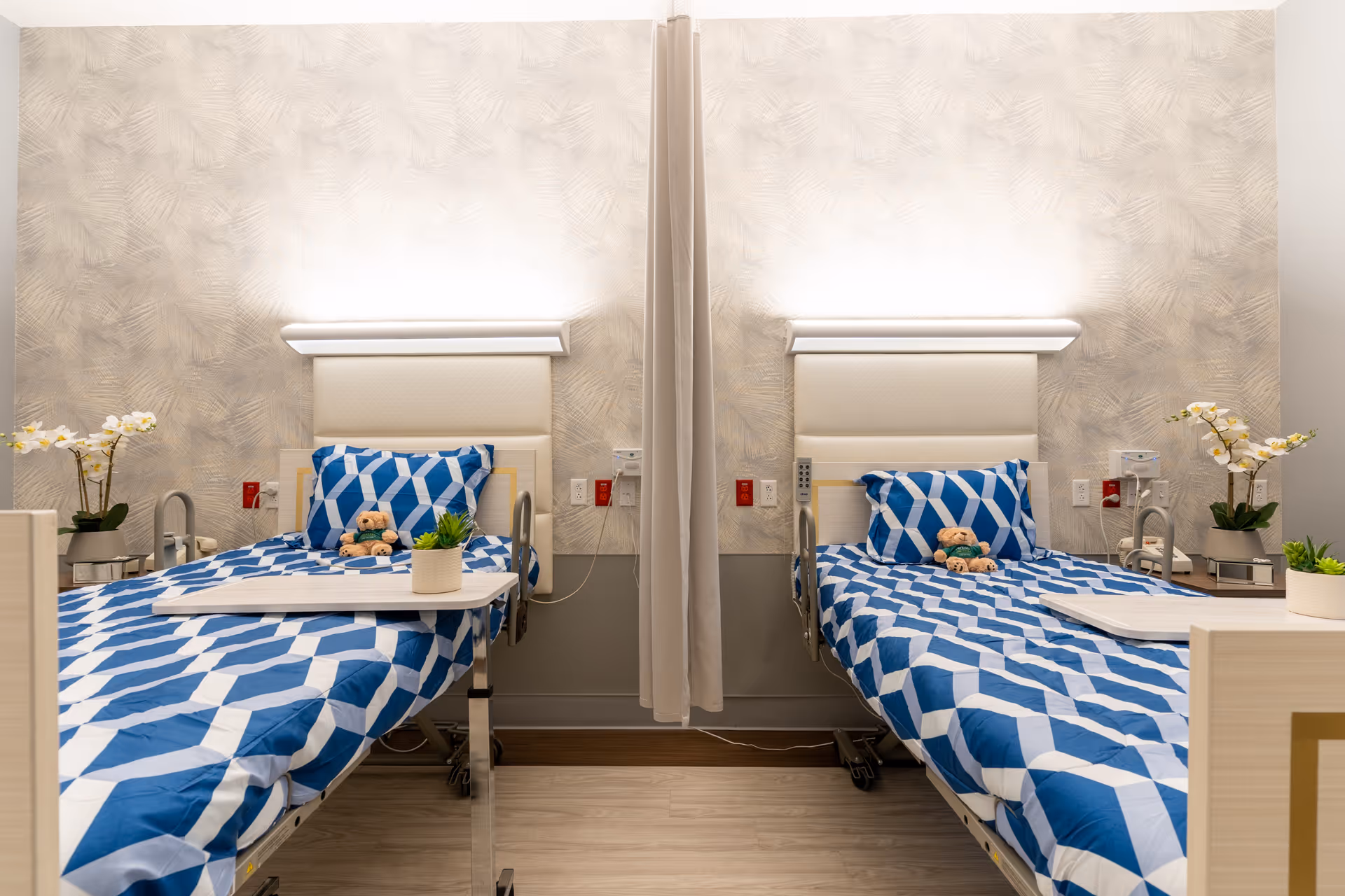 A clean and well-lit hospital or rehab room with two beds separated by a curtain. Each bed has blue and white geometric patterned bedding, a pillow, and a small teddy bear. There are bedside tables with potted plants and white orchids on each side. The walls have a textured beige design with soft lighting above the headboards.