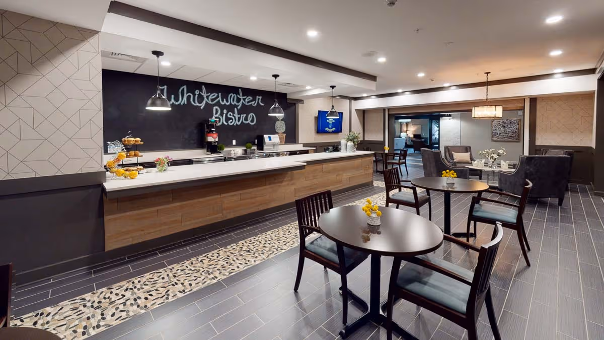 Interior view of a modern dining area named Whitewater Bistro with round tables and chairs, a long counter with pendant lights, and a seating area with armchairs and a coffee table in the background.