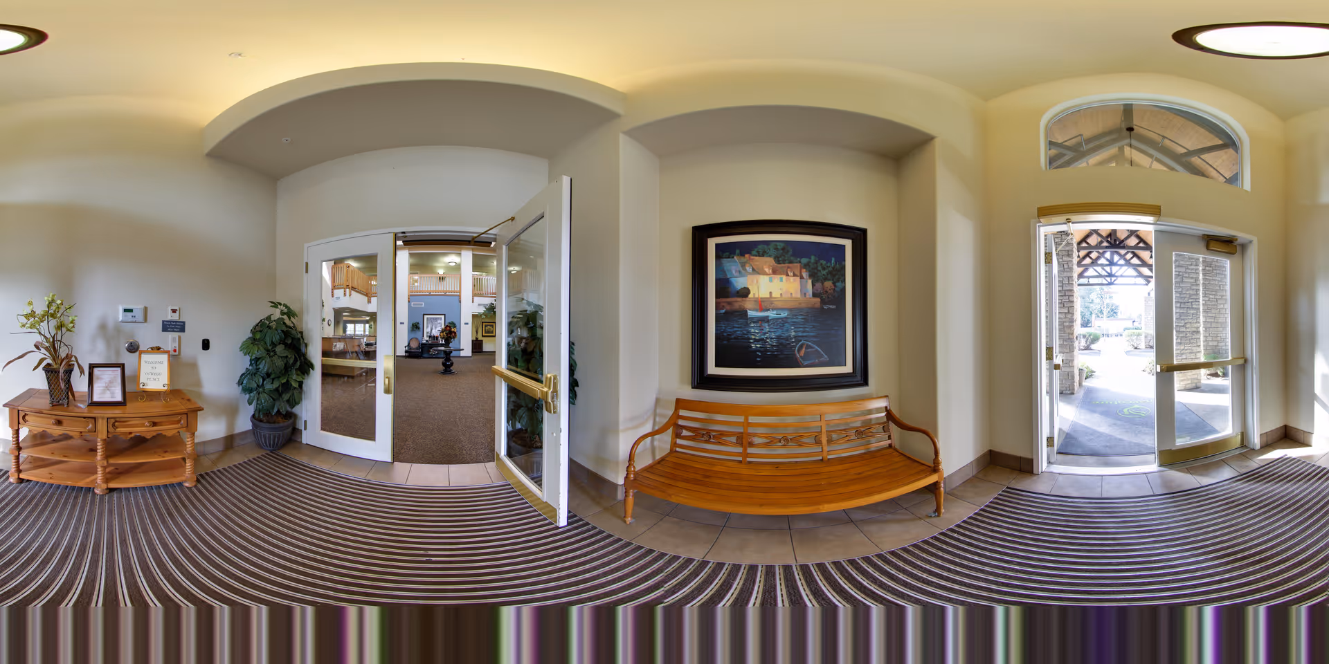 Entrance area of a senior living facility with double glass doors leading inside. There is a wooden bench under a framed painting on the right wall, and a wooden table with a plant and framed items on the left. The floor has a striped carpet and tile combination, and there is a large windowed door leading outside on the right.