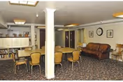 Interior view of a common area in a senior living facility featuring a long wooden table with several chairs around it, a brown leather sofa against the wall, framed pictures and a clock on the wall, and a bookshelf filled with books. The room has carpeted flooring and fluorescent ceiling lights.