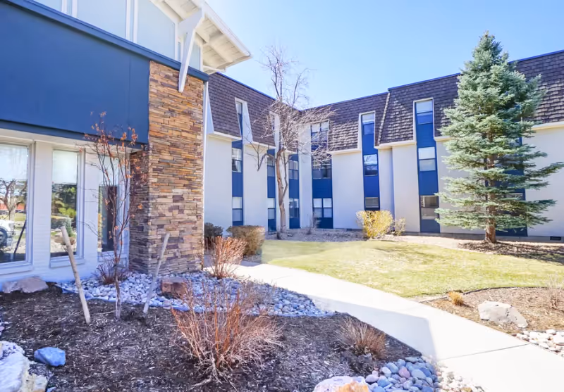 Exterior view of a senior living facility building with blue and white walls, multiple windows, a stone pillar, and landscaped grounds including bushes, a tree, and a grassy area under a clear blue sky.