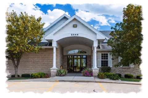 Front entrance of a one-story brick building with a covered portico displaying the number 5444, flanked by trees and landscaping.