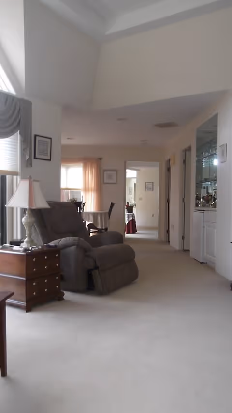 Interior view of a senior living facility showing a beige carpeted floor with a gray recliner chair next to a wooden side table with a lamp. In the background, there is a dining area with a table and chairs near a window with curtains. The hallway leads to other rooms, and there is a built-in glass cabinet on the right side.