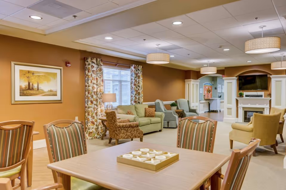 Communal living room/lounge with tables, upholstered chairs and sofas arranged around a fireplace and TV.