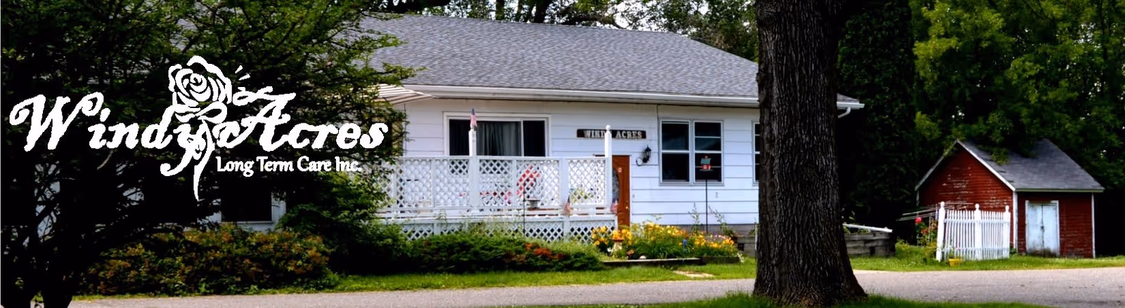 Exterior view of a single-story white building with a porch and railing, surrounded by trees and greenery. There is a small red shed with a white door and fence to the right. The building has a sign that reads 'Windy Acres'.