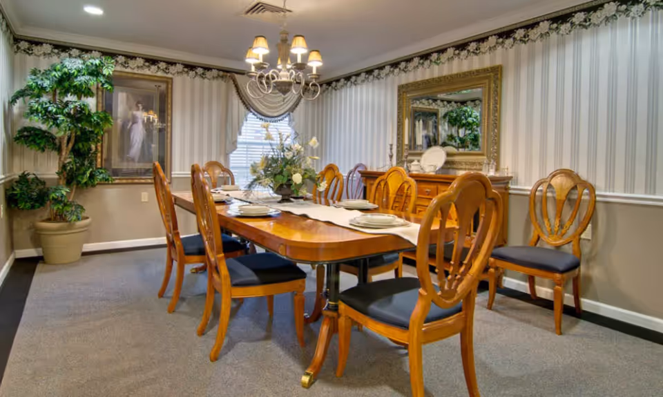 A formal dining room with a long wooden table set with plates and a floral centerpiece. The room features eight wooden chairs with dark cushions, a large mirror on the wall, a chandelier hanging from the ceiling, striped wallpaper, and a potted plant in the corner.