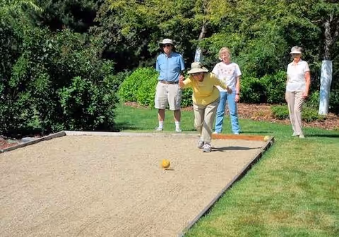 Four seniors playing bocce ball outdoors on a bocce court surrounded by green grass and trees. One woman is in the act of rolling a bocce ball while three others watch.