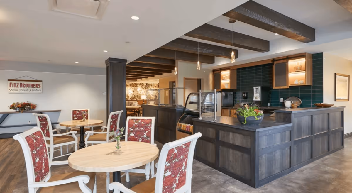 Communal dining area with round wooden tables and upholstered chairs in front of a dark wood service counter and exposed ceiling beams.