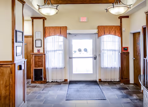Bright lobby entry with a glass door flanked by curtained windows, tile flooring, wood trim, and hanging ceiling lights.