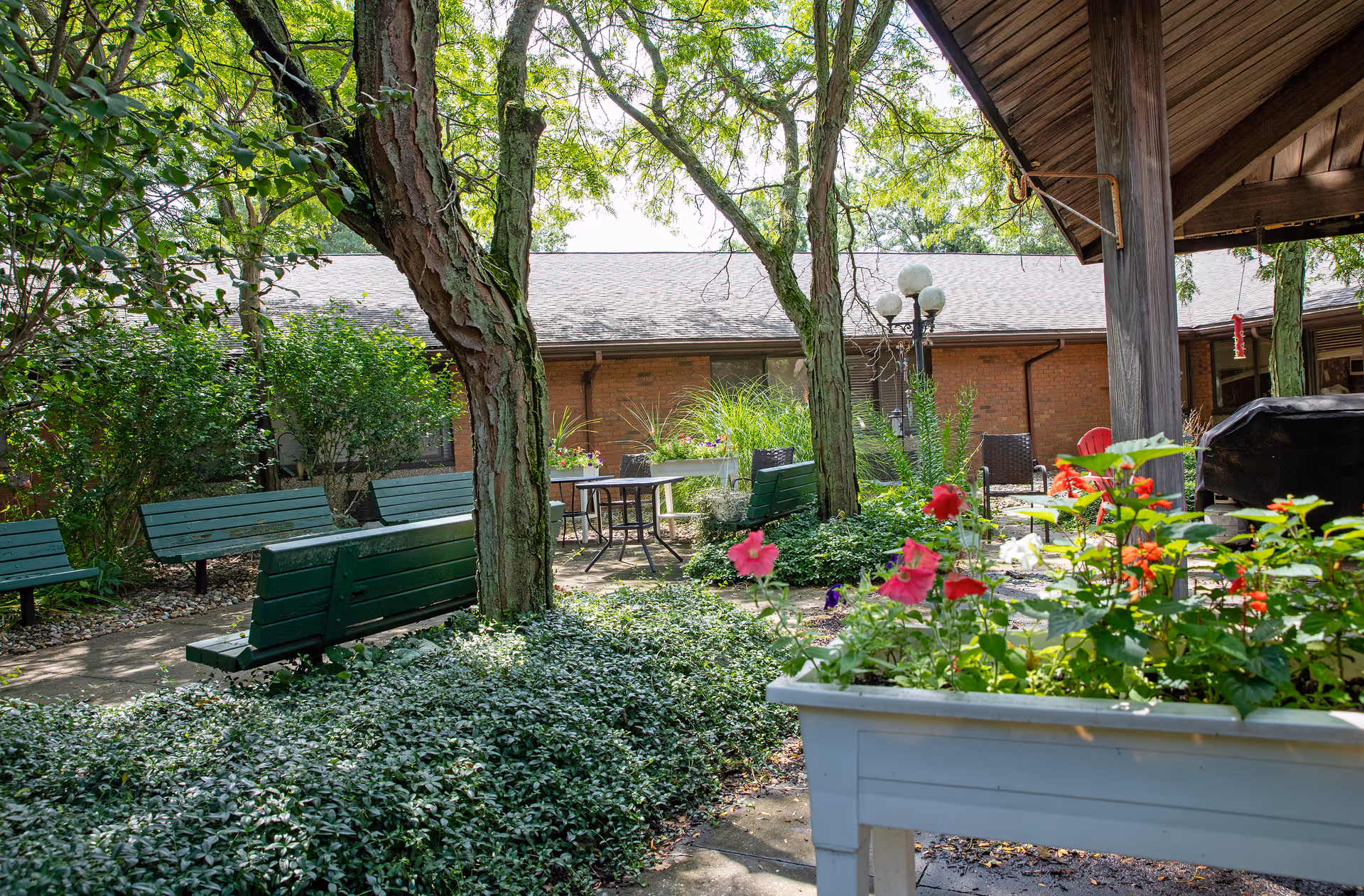 A peaceful outdoor garden area at East Lake Nursing and Rehabilitation Center featuring green benches, trees, flowering plants, and a covered patio with seating and a grill.