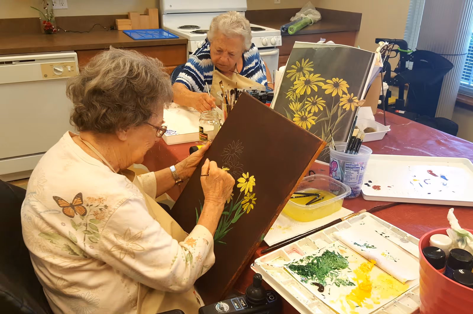 Two elderly women seated at a table in a kitchen area, engaged in painting yellow flowers on canvases. The table is covered with art supplies including paint palettes, brushes, and jars. The background shows kitchen appliances and a window with blinds.