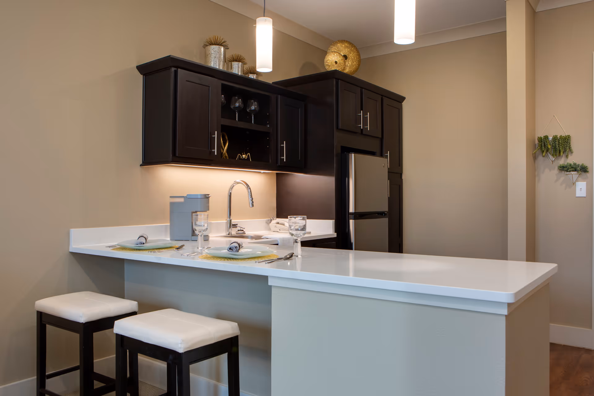 Modern kitchen area with dark wood cabinets, a stainless steel refrigerator, a white countertop island with two cushioned stools, and place settings with plates, napkins, and glasses. Decorative items are placed on top of the cabinets and a small plant hangs on the wall.