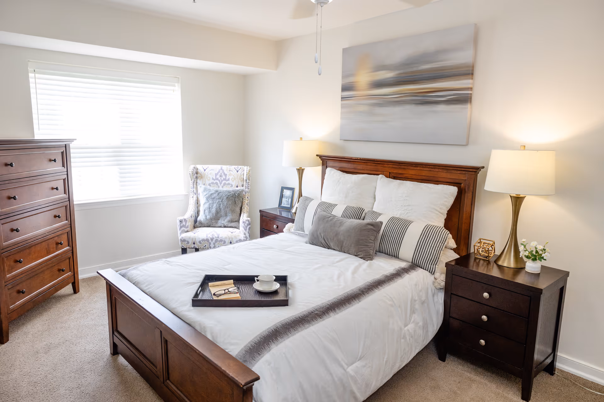 A bright and cozy bedroom featuring a wooden bed with white and gray bedding, two bedside tables each with a lamp, a wooden dresser, a patterned armchair with a gray pillow, and a large window with blinds letting in natural light. A large abstract painting hangs above the bed.