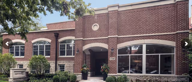 Exterior view of a brick building with large windows and an arched entrance, surrounded by greenery and trees. A sign near the entrance reads 'aperion care'.