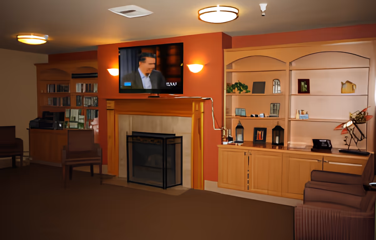 A cozy living room area in an assisted living facility featuring a wall-mounted flat-screen TV above a fireplace with a wooden mantel. The walls are painted a warm orange color. On either side of the fireplace are built-in wooden shelves and cabinets displaying decorative items, books, and framed pictures. There are several chairs arranged around the room, and the lighting includes ceiling fixtures and wall sconces.