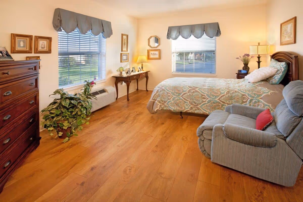 A cozy bedroom with wooden flooring, a bed with a patterned bedspread, a gray recliner chair with a red pillow, a wooden dresser, a small table with framed photos and a lamp, two windows with blue valances, and a potted plant near the window.