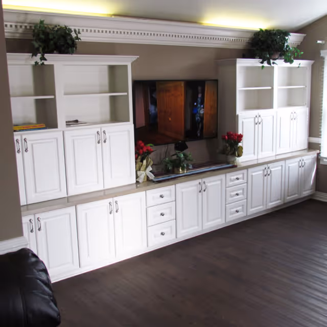 Living room wall with white built-in cabinets and shelves, a mounted TV, and decorative plants and flowers.