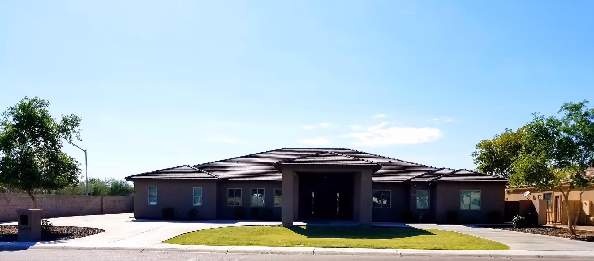 Front exterior view of Quail Manor Assisted Living facility, a single-story building with a tiled roof, surrounded by a lawn and trees under a clear blue sky.