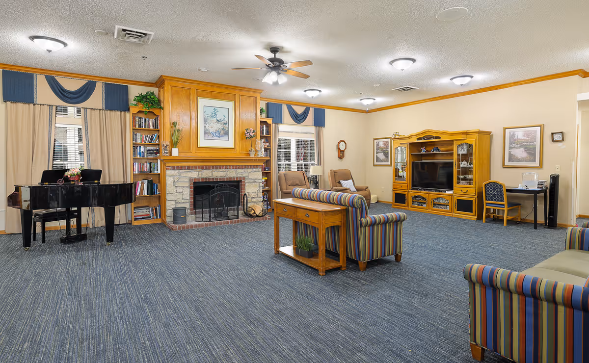 A spacious living room in an assisted living facility featuring a stone fireplace with a wooden mantle, flanked by built-in bookshelves. The room has multiple seating options including striped sofas and armchairs, a black grand piano near the window, a wooden entertainment center with a TV, and framed artwork on the walls. The carpet is blue, and the ceiling has multiple light fixtures and a ceiling fan.