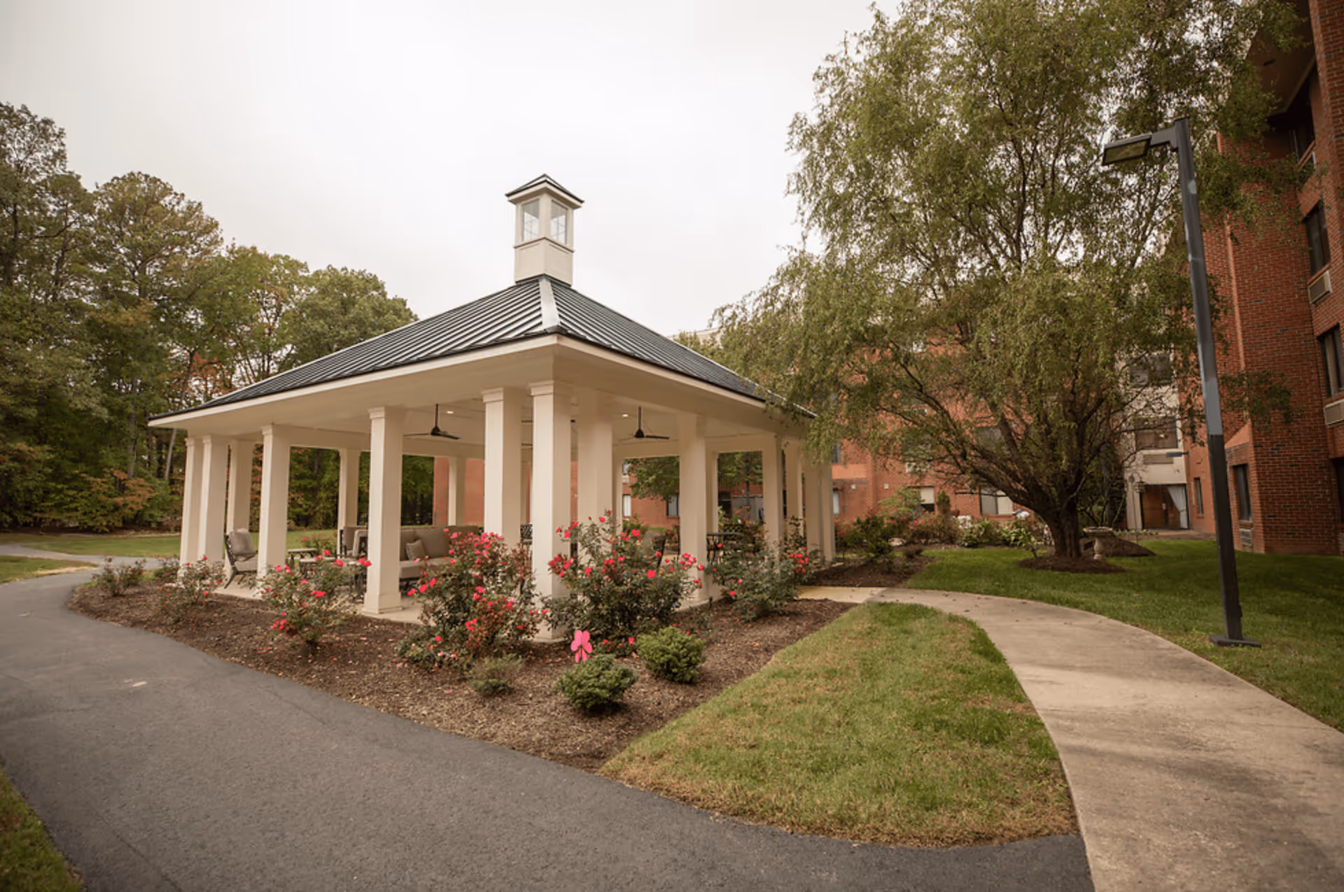 Outdoor gazebo structure with white pillars and a dark roof, surrounded by flower beds with pink flowers and greenery. There are chairs and tables under the gazebo. A paved pathway curves around the gazebo, and a large tree and a brick building are visible in the background.