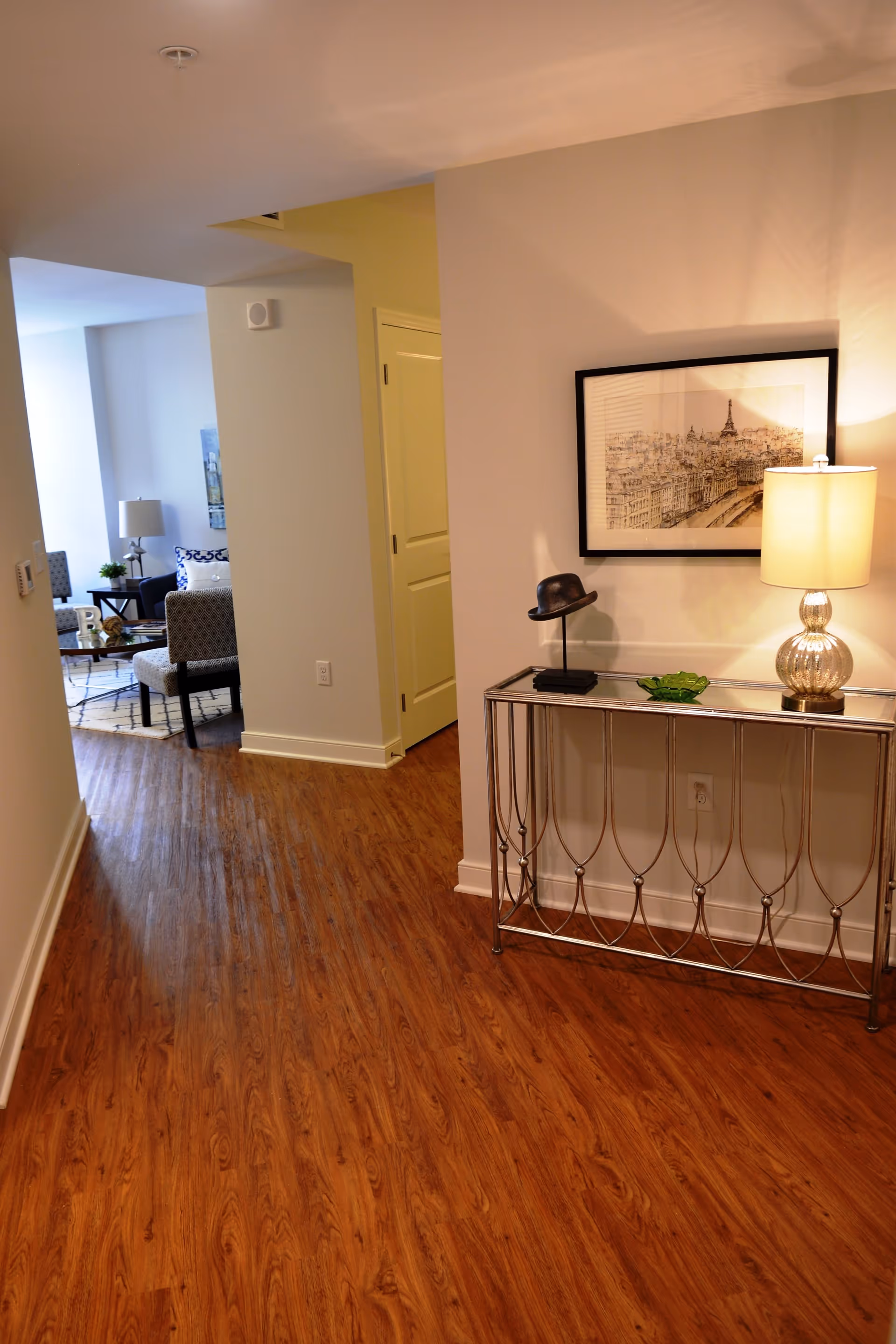 Hallway with wood-look flooring leading into a living area, featuring a console table with a lamp and framed artwork on the wall.