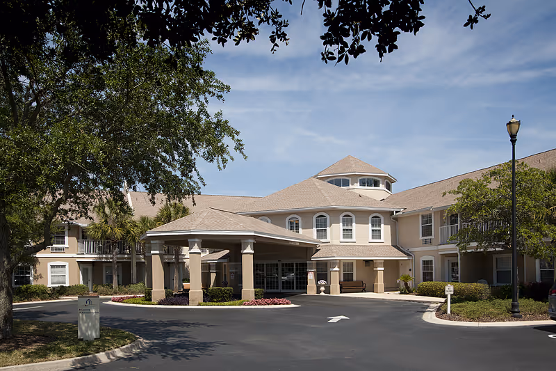 Exterior view of Allegro Senior Living facility in Fleming Island, FL, showing a two-story building with beige walls and a covered entrance. The building is surrounded by trees, shrubs, and a paved driveway with a streetlamp on the right side under a clear blue sky.