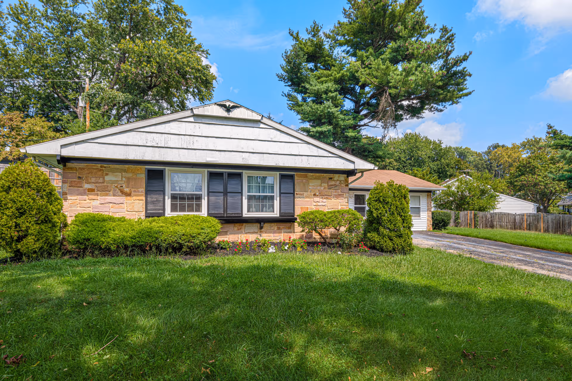 Single-story residential building with a stone facade and white siding on the upper part, black shutters on the windows, surrounded by green bushes and a well-maintained lawn under a blue sky with some clouds.