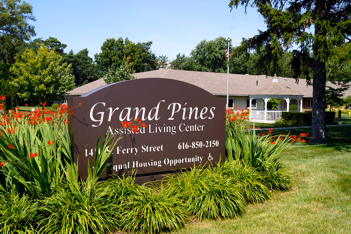Outdoor view of Grand Pines Assisted Living Center sign surrounded by green plants and red flowers, with the facility building and trees in the background under a clear blue sky.
