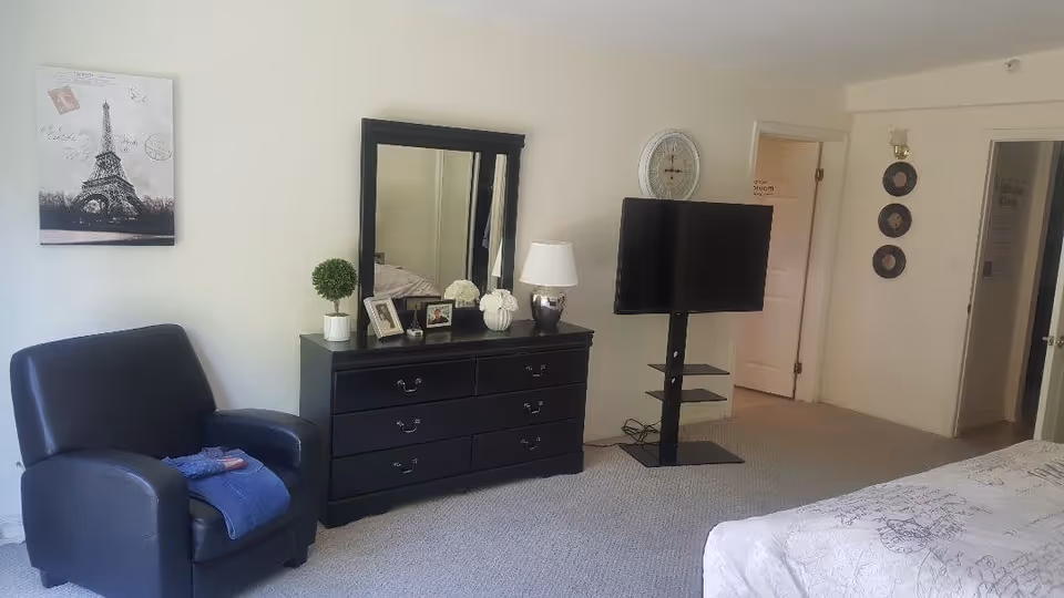 A senior living bedroom featuring a black armchair with a folded blue towel on it, a black dresser with a large mirror, framed photos, a small potted plant, a lamp, and a vase with white flowers. A flat-screen TV is mounted on a stand next to the dresser. The room has light-colored walls, carpeted floor, and a bed partially visible on the right side. There is a wall clock above the TV and decorative plates on the wall near the doorways.