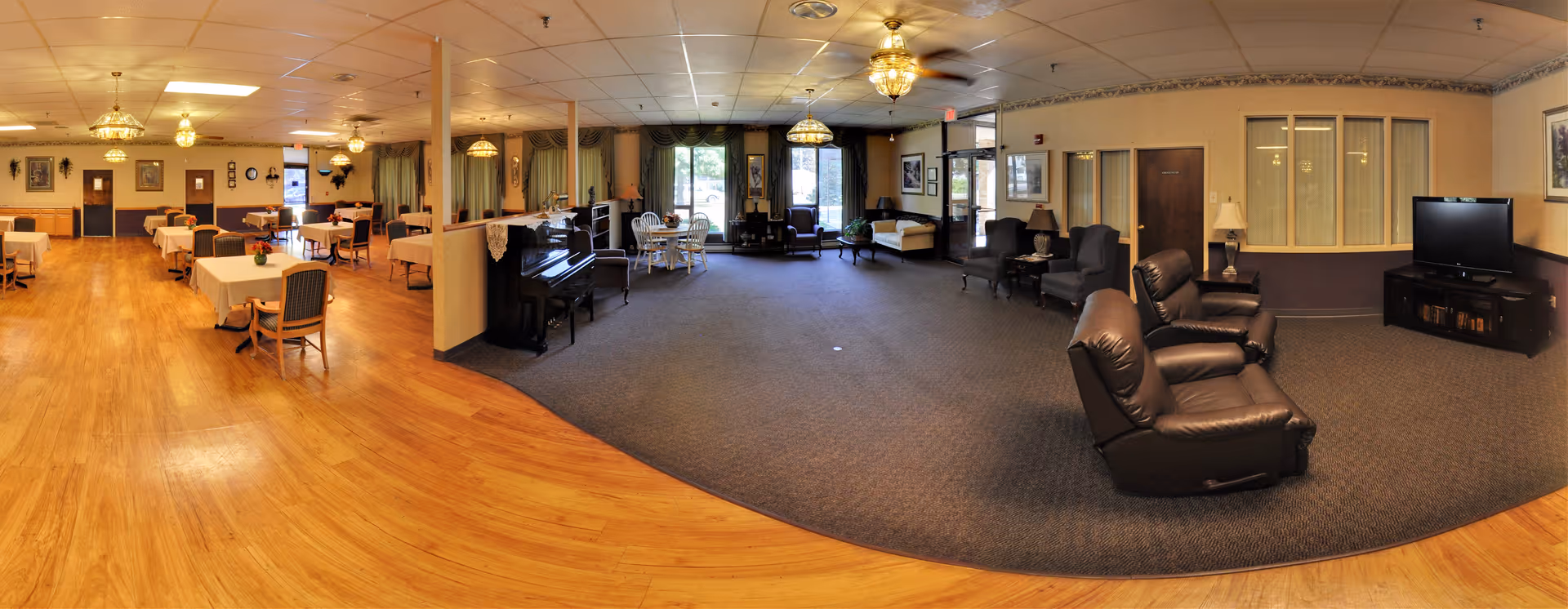 Communal senior living common room with dining tables on hardwood floors to the left and a carpeted lounge area with recliners, piano, and TV to the right.