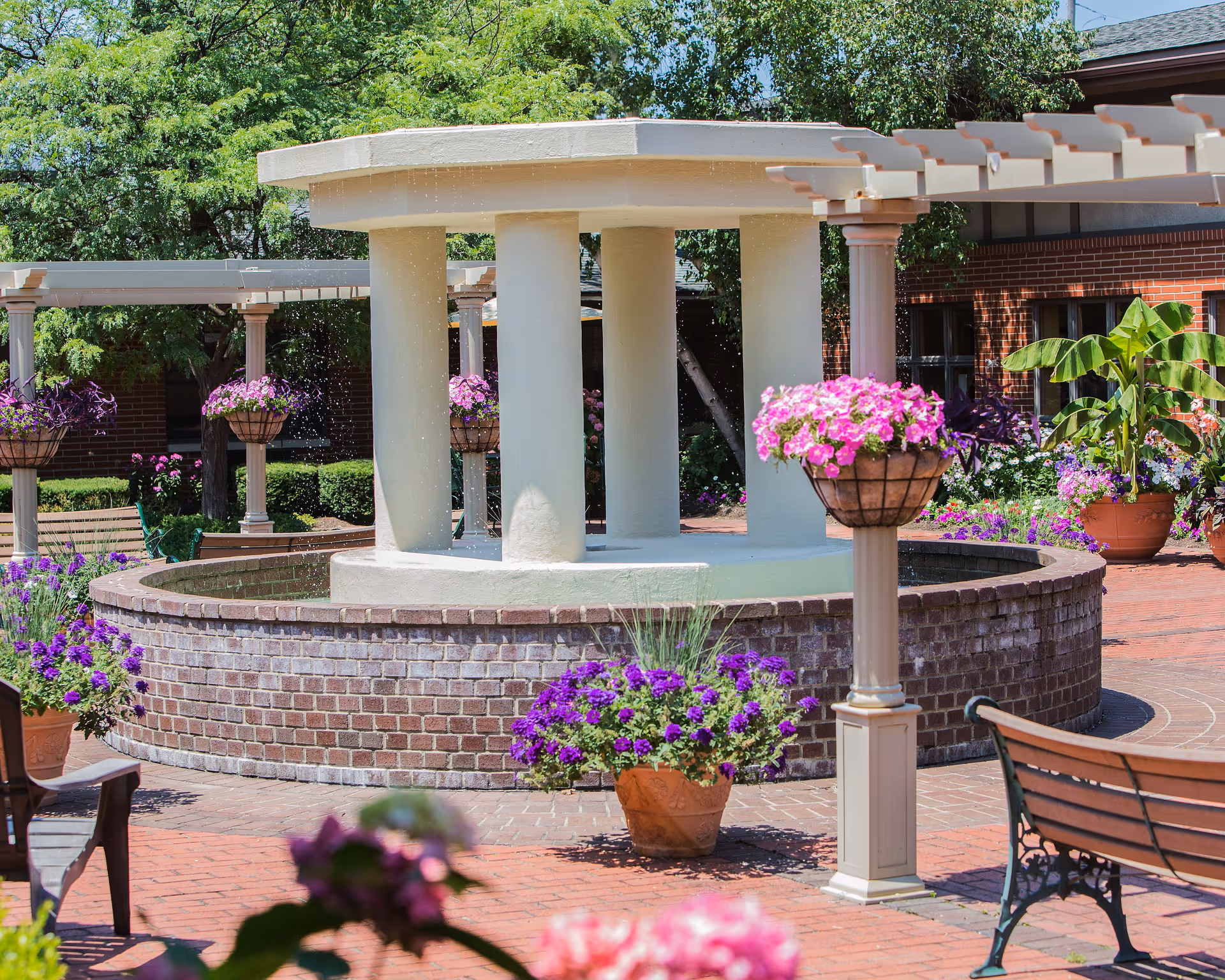 Outdoor courtyard area with a circular brick fountain surrounded by hanging baskets and pots of purple and pink flowers, benches, and a pergola structure with greenery and trees in the background.