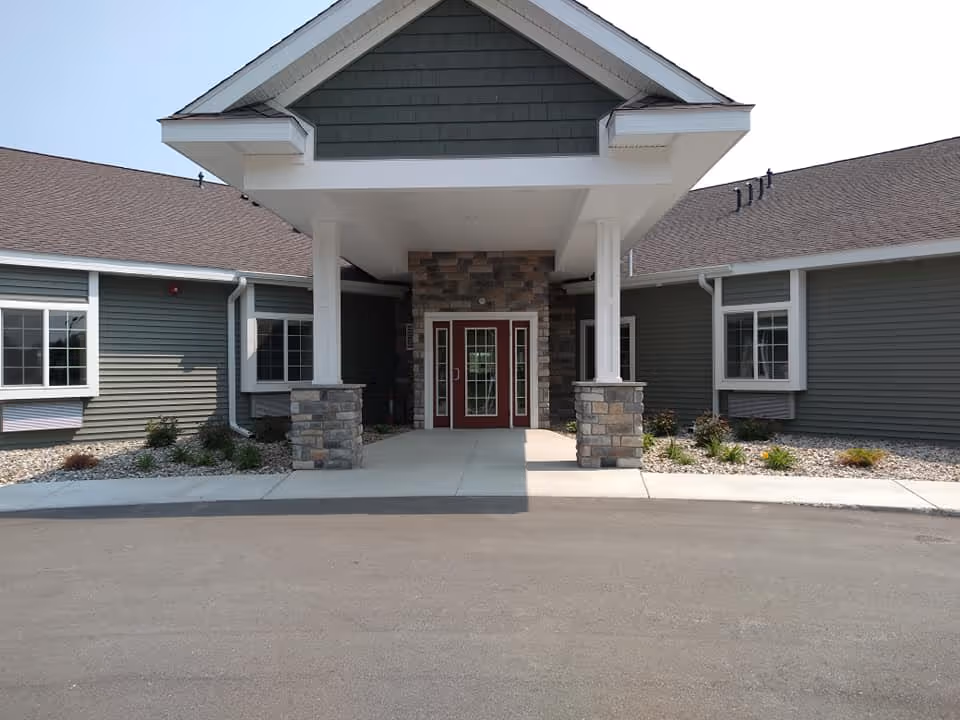 Front exterior view of Coleman Fields Assisted Living & Memory Care building with a covered entrance supported by two columns with stone bases, gray siding, and a peaked roof.