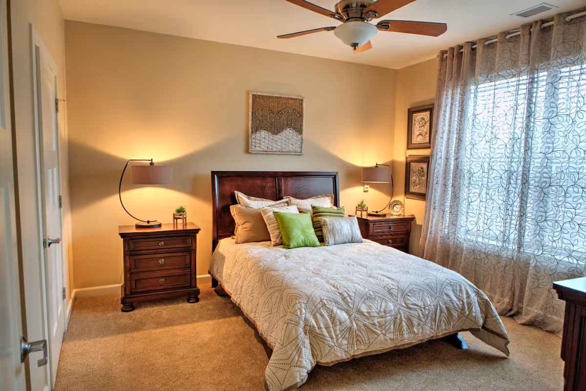 Warm, well-lit bedroom with a bed, two wooden nightstands, bedside lamps, ceiling fan and curtained window.