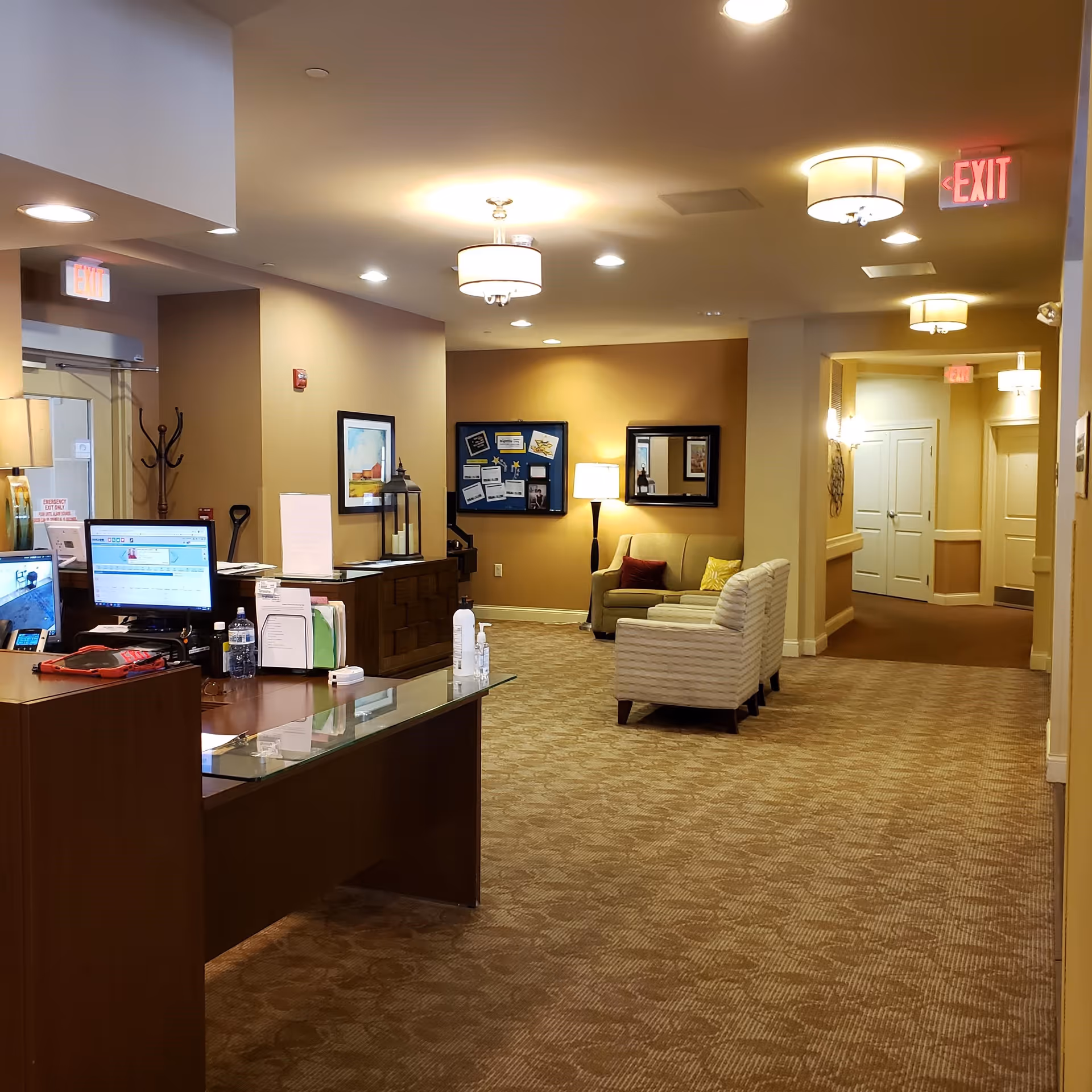 Interior view of a senior living facility reception area with a wooden desk equipped with computer monitors, office supplies, and a water bottle. The space has beige walls and carpeted floors, with a seating area featuring a sofa and two armchairs near a floor lamp and a bulletin board. Multiple ceiling lights and exit signs are visible, along with a hallway leading to closed doors.