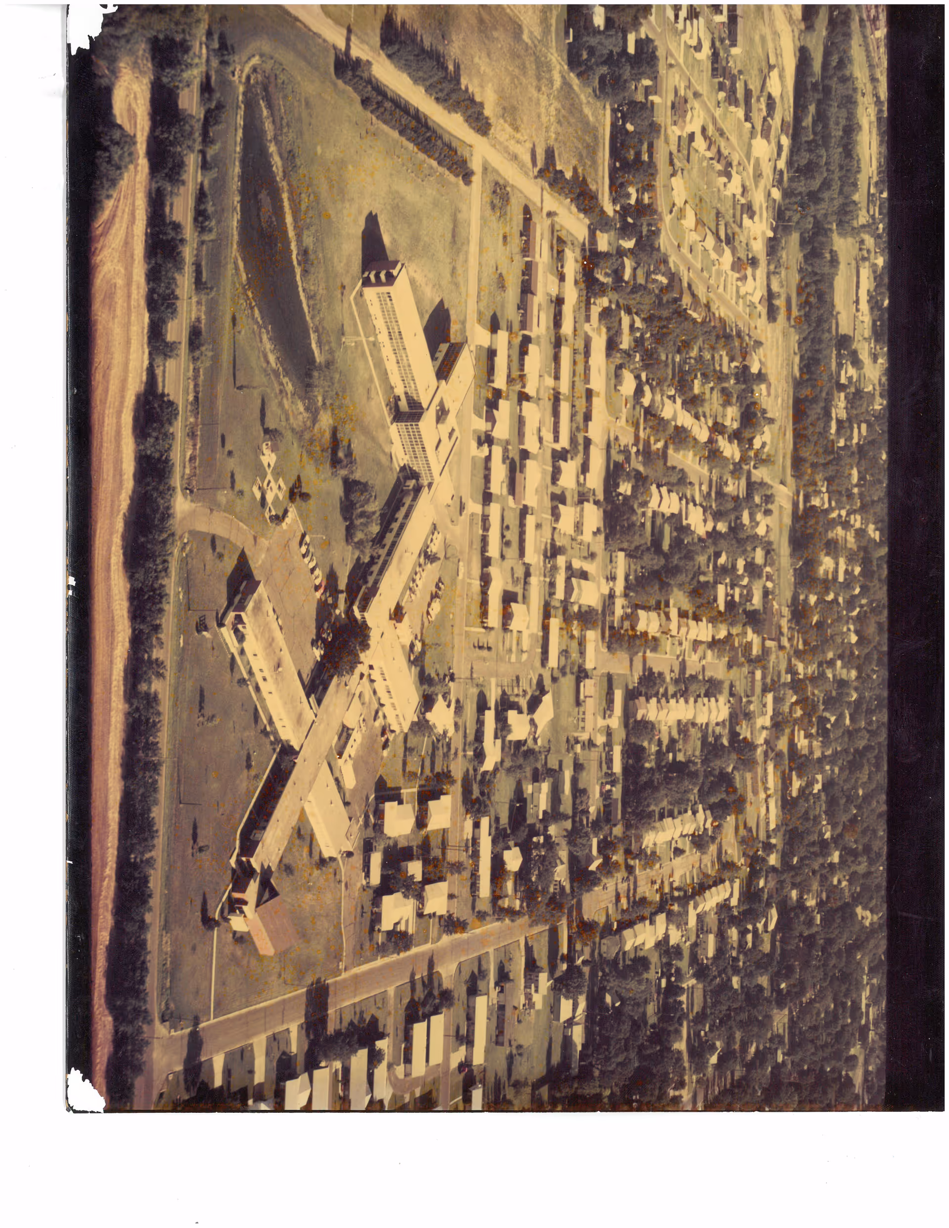 Aerial view of a large building complex with multiple wings surrounded by a residential neighborhood with many houses and streets. The complex is situated near a body of water and open green spaces.