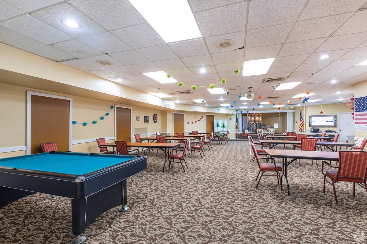 A large community room with patterned carpet, multiple rectangular tables with red cushioned chairs, and a pool table with a blue felt top. The ceiling has fluorescent lights and decorative hanging pom-poms in various colors. The walls are beige with some decorations and an American flag is visible near a television screen mounted on the far wall.