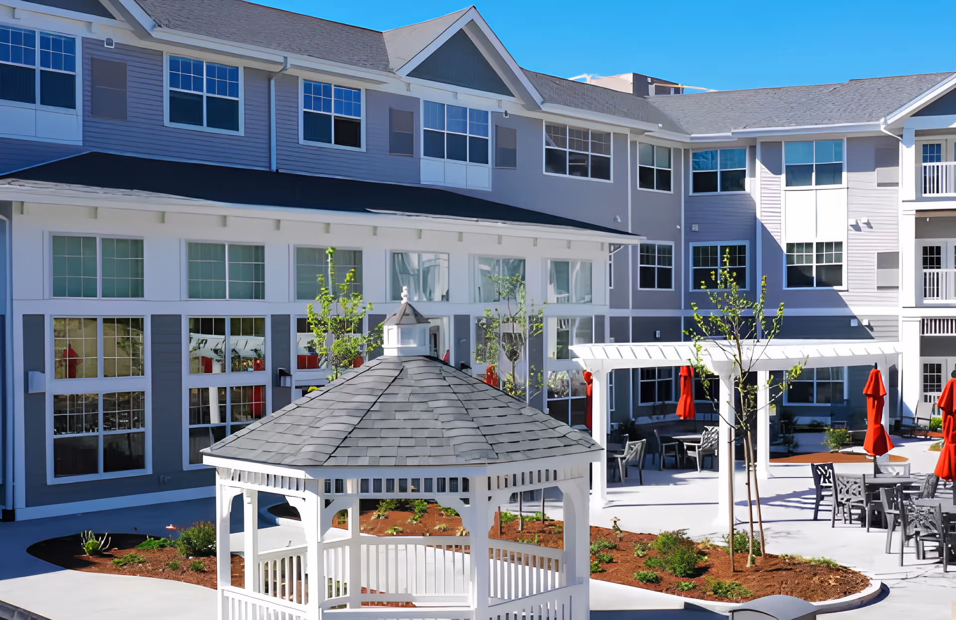 Outdoor courtyard area of The Woods - Retirement Community featuring a white gazebo in the foreground, surrounded by landscaped garden beds, young trees, and patio seating with tables and red umbrellas. The multi-story building with large windows forms the background under a clear blue sky.