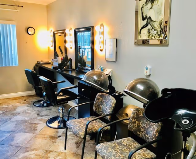 Interior view of a salon area with three black salon chairs in front of mirrors mounted on the wall, two floral patterned chairs, two hair drying machines, and a black hair washing station. The room has tiled flooring, a clock on the wall, and soft lighting from wall-mounted fixtures.