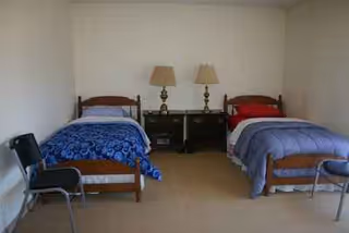 Two single beds with nightstands and lamps in a plain, shared bedroom.