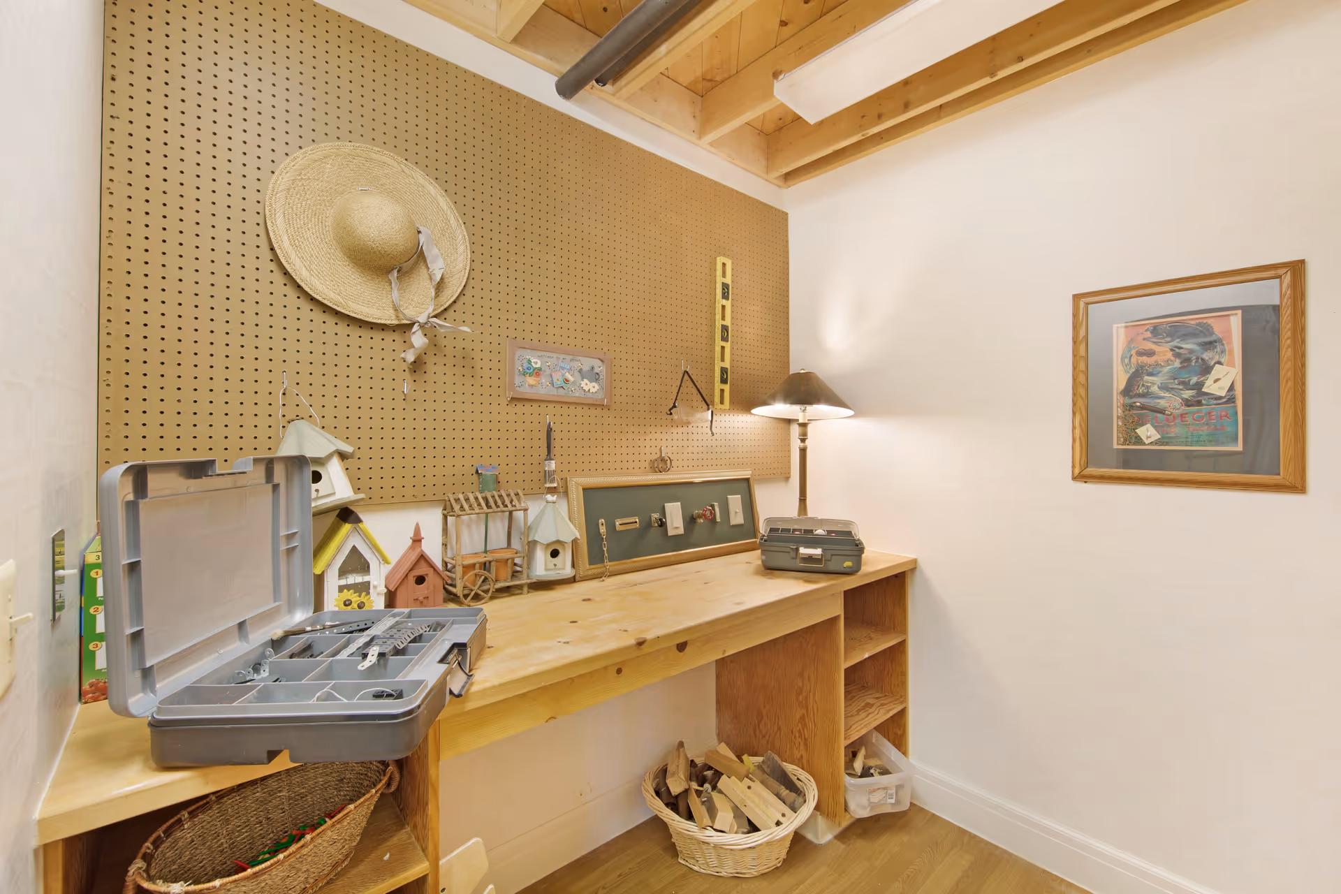 Small craft/workbench room with a wooden counter, pegboard holding a straw hat and small birdhouses, toolboxes and baskets beneath the counter.