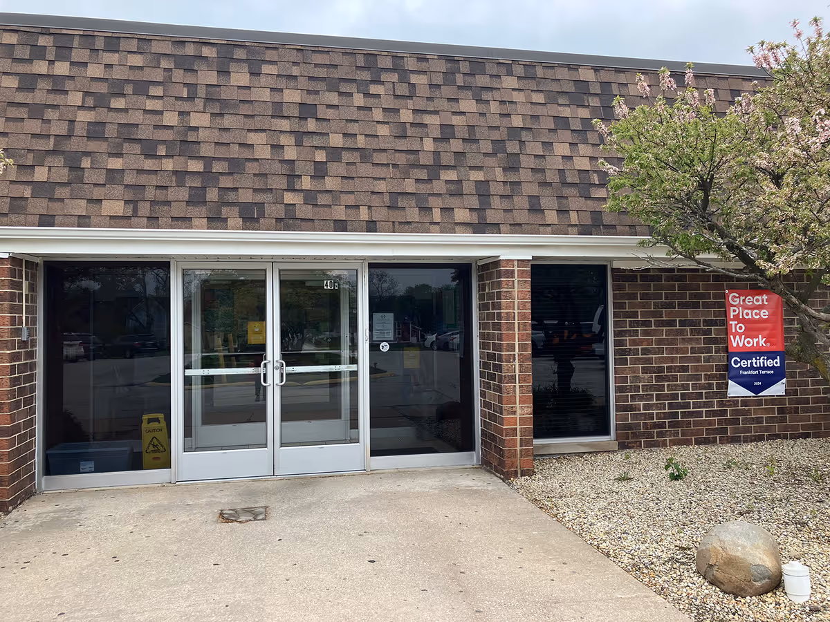 Exterior view of a brick building entrance with double glass doors and windows on either side. There is a tree with pink blossoms on the right side and a sign on the wall that reads 'Great Place To Work Certified Frankfort Terrace 2024'.