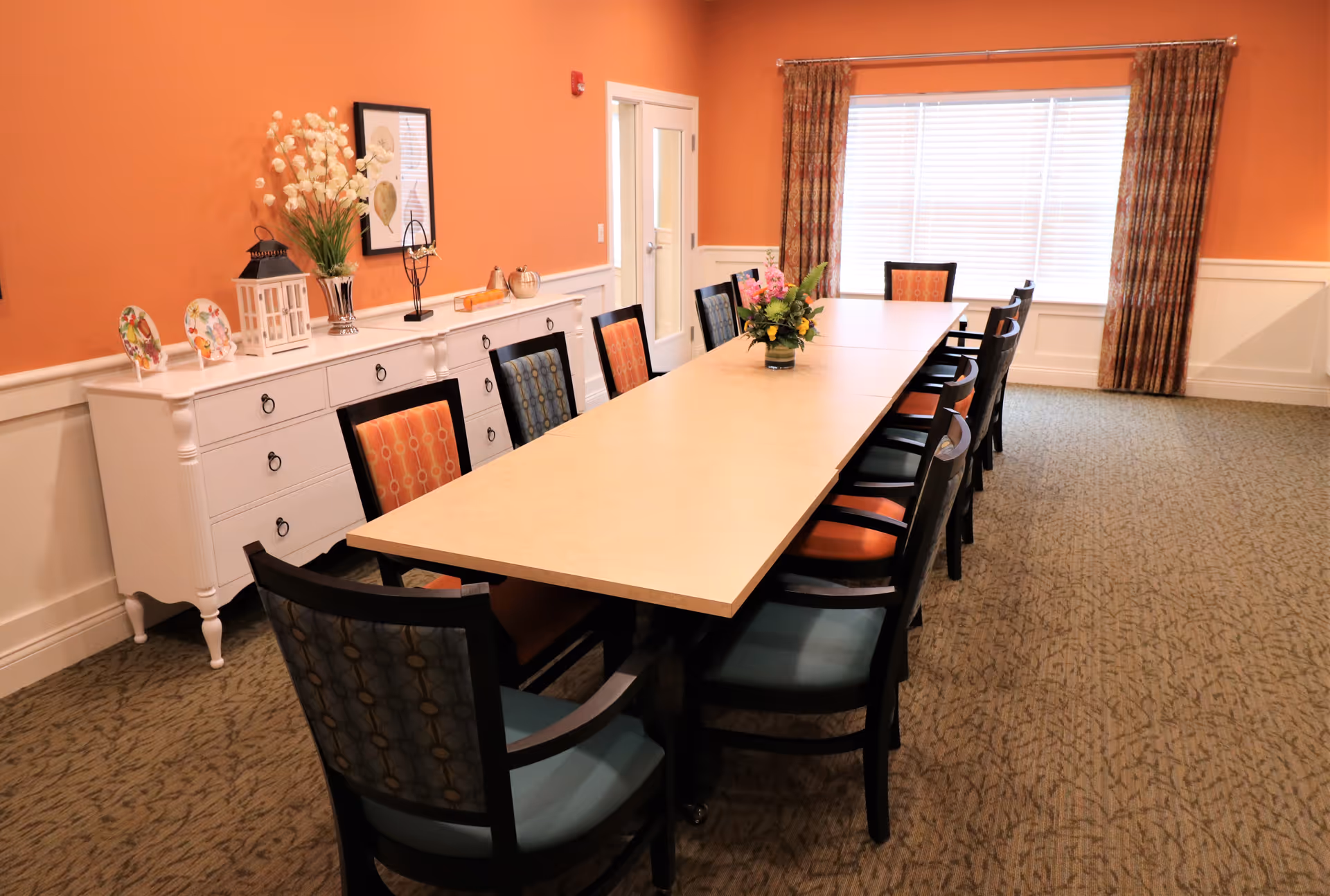 A dining room with a long rectangular table surrounded by chairs with patterned upholstery in orange and blue. The walls are painted orange with white wainscoting, and there is a large window with patterned curtains letting in natural light. A white sideboard against the wall holds decorative items including plates, a lantern, a vase with flowers, and framed artwork.