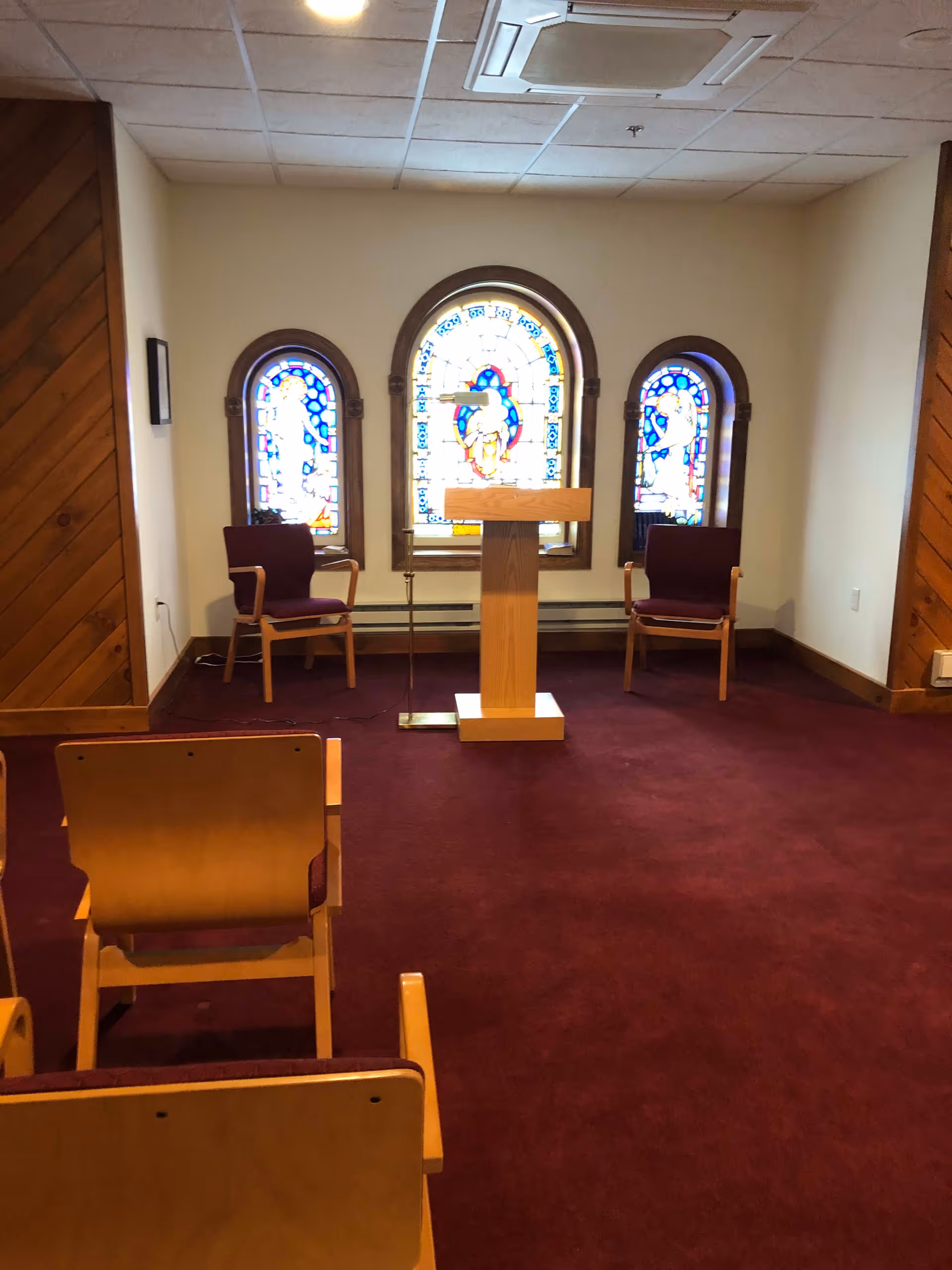 Small chapel or prayer room with three stained glass windows on the far wall. There is a wooden podium in the center with a microphone stand next to it. Two wooden chairs with purple cushions are placed on either side of the podium. Several wooden chairs with purple cushions are arranged facing the podium on a maroon carpeted floor. The walls are light-colored with wood paneling on the left and right edges.