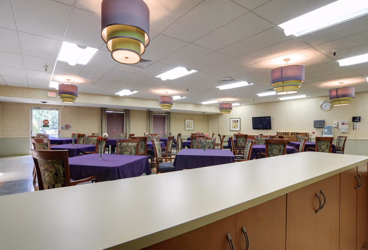 Interior view of a dining room in Charleston Healthcare Center with multiple tables covered in purple tablecloths, each adorned with a small vase of flowers. The room has patterned wallpaper, several decorative ceiling lights, a wall-mounted TV, and a clock. There are wooden chairs with patterned cushions around the tables.