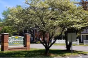 Outdoor view of the entrance to Westbrook Towers senior living facility, featuring a green sign with white text mounted on a brick base, surrounded by trees and greenery.