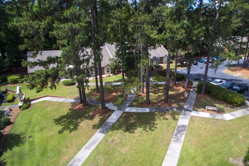 Aerial view of Savannah Square grounds with intersecting walkways, tall pine trees, grassy lawns, nearby residential buildings, and a parking area.