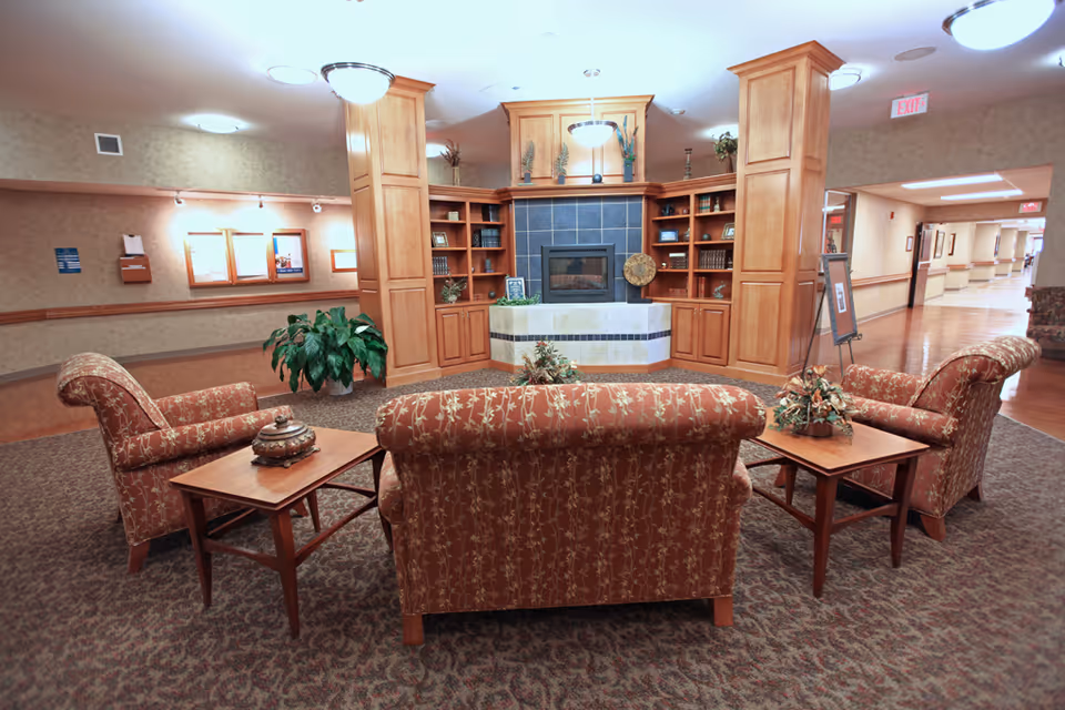 A cozy common area with patterned upholstered chairs and a loveseat arranged around two wooden side tables. In the background, there is a built-in wooden shelving unit with decorative items and a fireplace in the center. The room has carpeted flooring and warm lighting, with a hallway visible to the right.