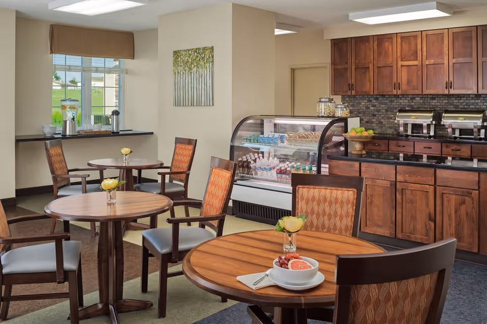 A cozy dining area with round wooden tables and cushioned chairs. Each table has a small vase with a yellow flower. In the background, there is a counter with wooden cabinets, a refrigerated display case with bottled drinks and snacks, and two food warmers. A window with a brown valance lets in natural light, and a painting of trees hangs on the wall.