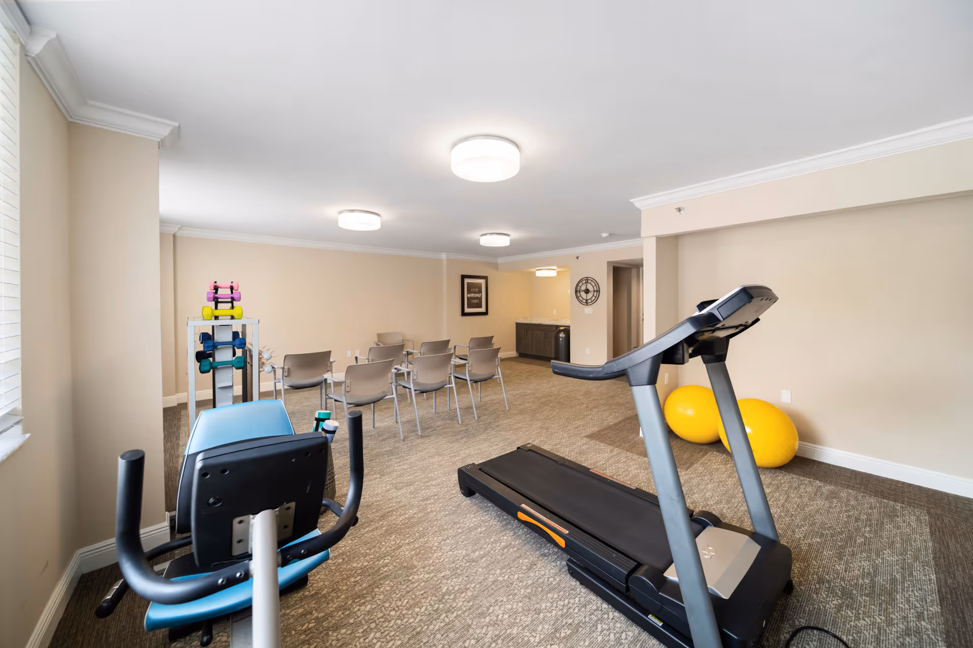A fitness room in a senior living facility with exercise equipment including a treadmill, a recumbent bike, colorful dumbbells on a rack, and two large yellow exercise balls. There are several chairs arranged in rows facing a wall with a framed sign and a clock. The room has beige walls, carpeted floor, and ceiling lights.